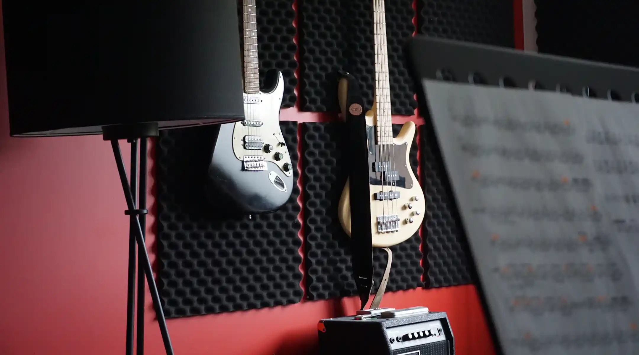 A music studio wall with red paint and acoustic foam panels featuring a black electric guitar and a cream bass guitar hanging above an amplifier