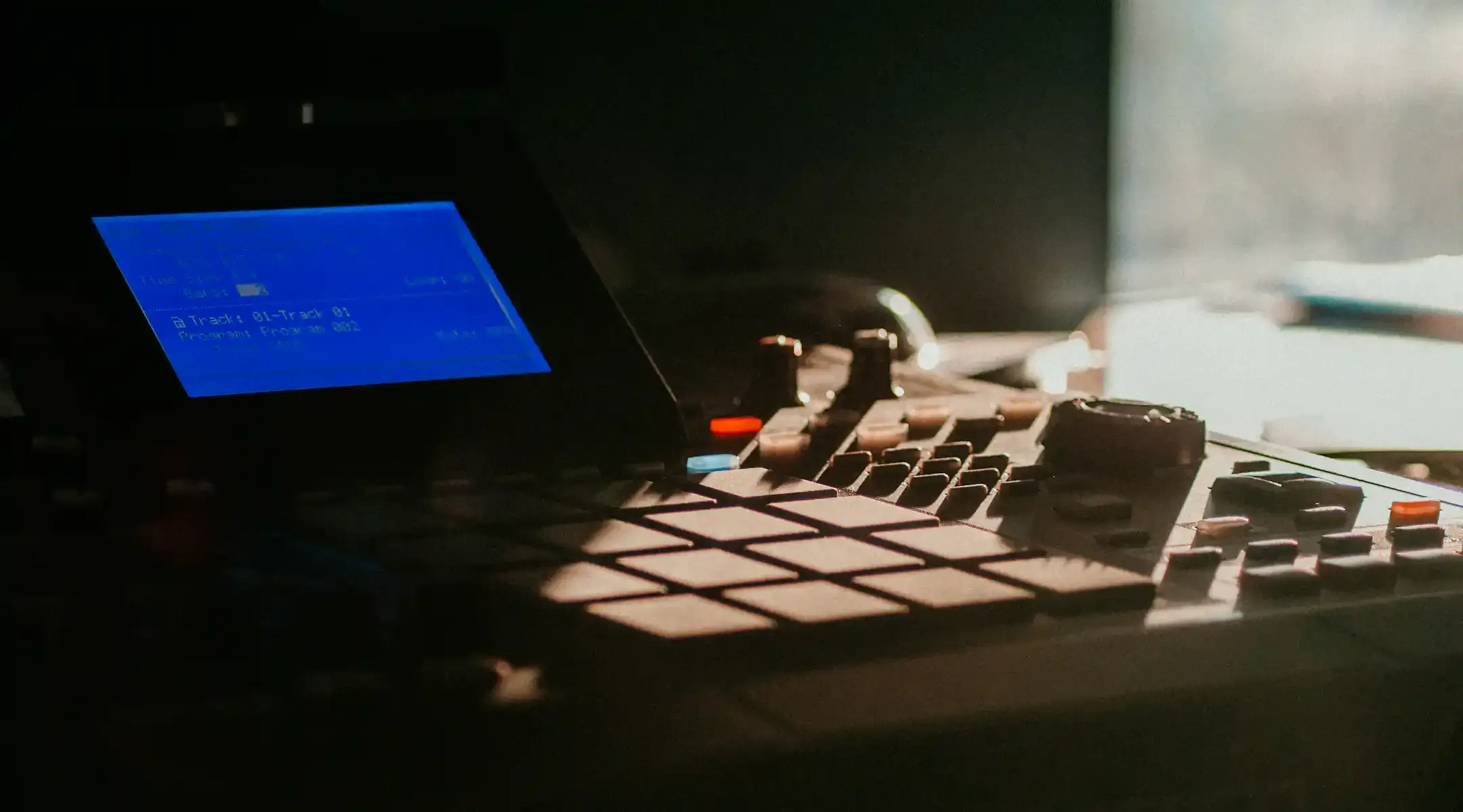 Close-up of a music production device with a blue screen displaying track information and various buttons, set in dim lighting