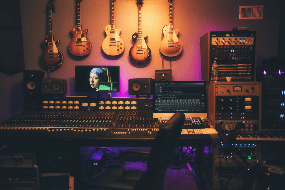 A music studio with an array of guitars on the wall, mixing consoles, monitors, and sound equipment, lit with purple hues.
