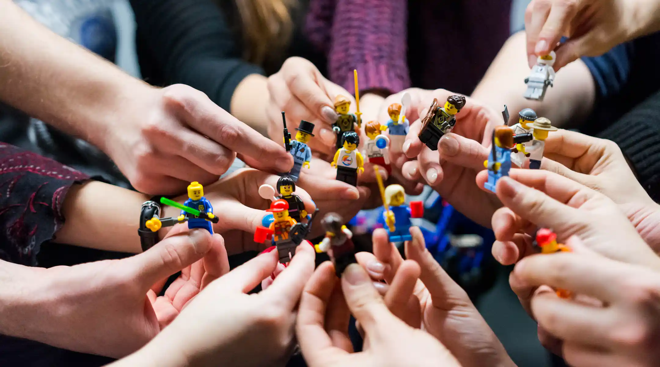 Multiple hands holding up various colorful LEGO minifigures in a circle during a group activity