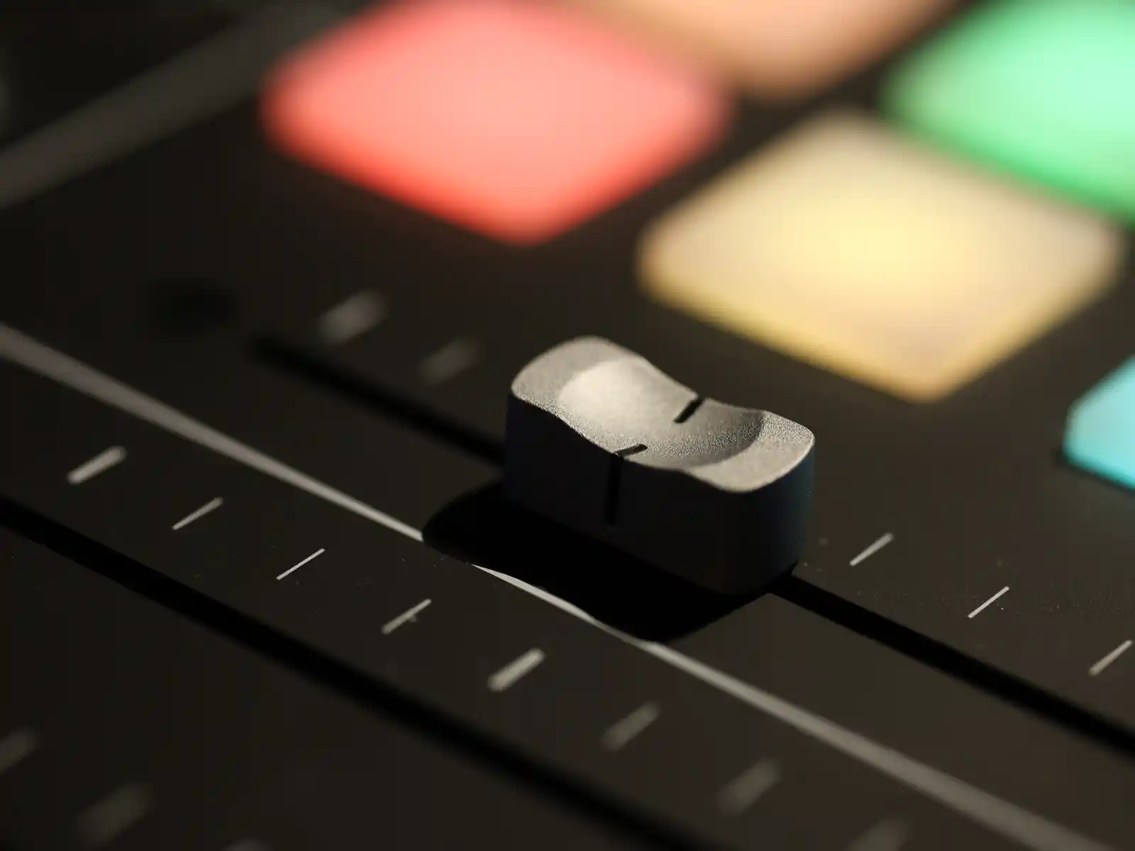 A macro shot of a single black fader on a MIDI controller with colorful backlit pads in the background